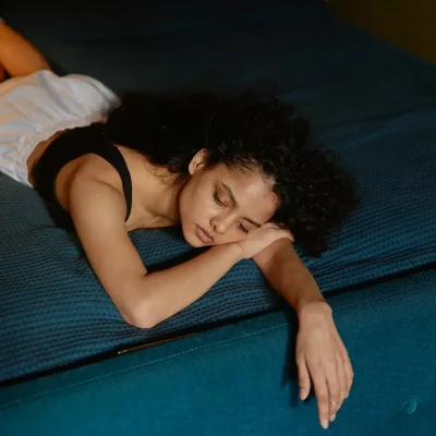 Tired woman lying on a bed with eyes closed and arm stretched out, resting on a blue blanket.
