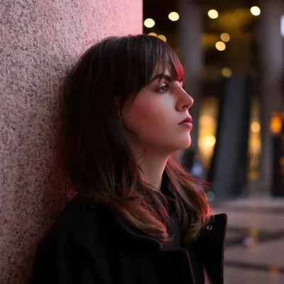 A woman with a serious expression, leaning on a wall with city lights blurred in the background, evoking contemplation or concern.
